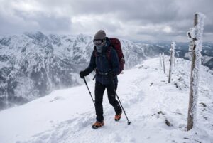 Hiker in winter layers, goggles and microspikes walking with trekking poles along a snowy mountain ridge