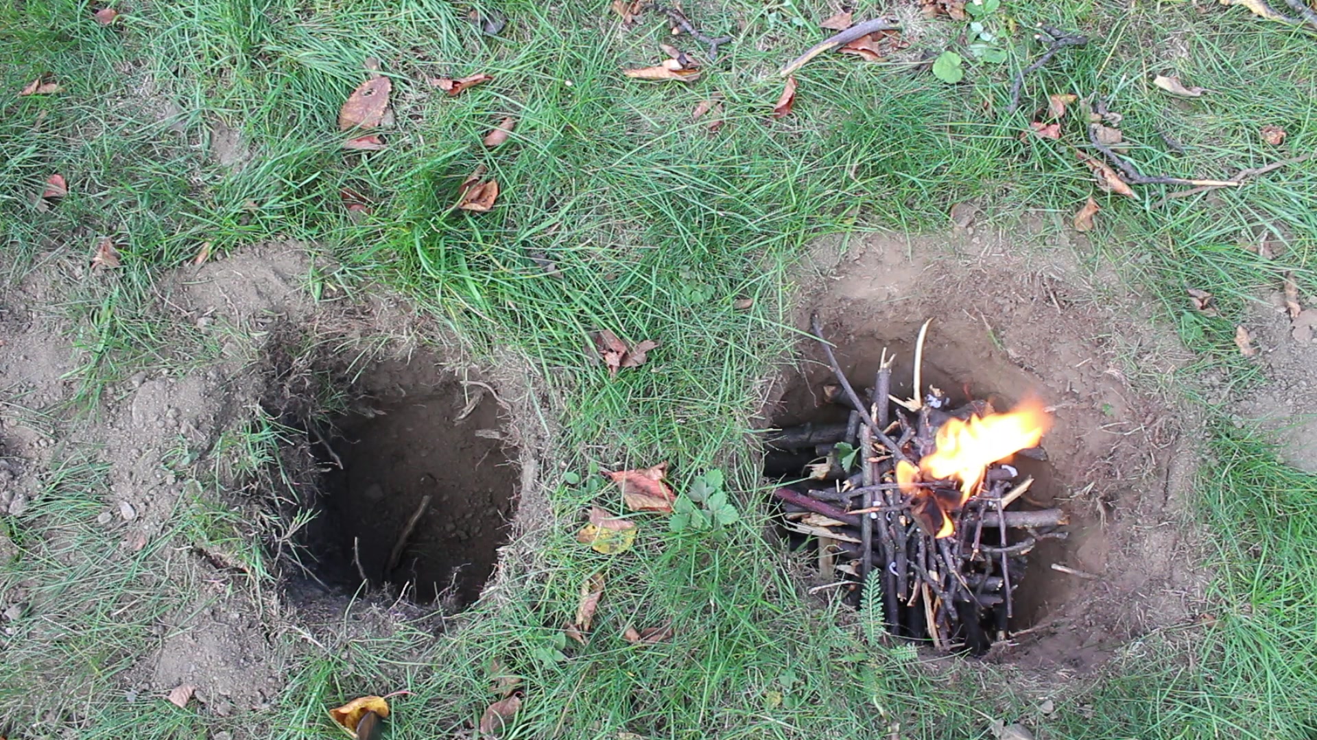 Lighting a Dakota fire, sticks laid crosswise in layers in the pit, a flame on one side of the stack, the empty air channel next to it