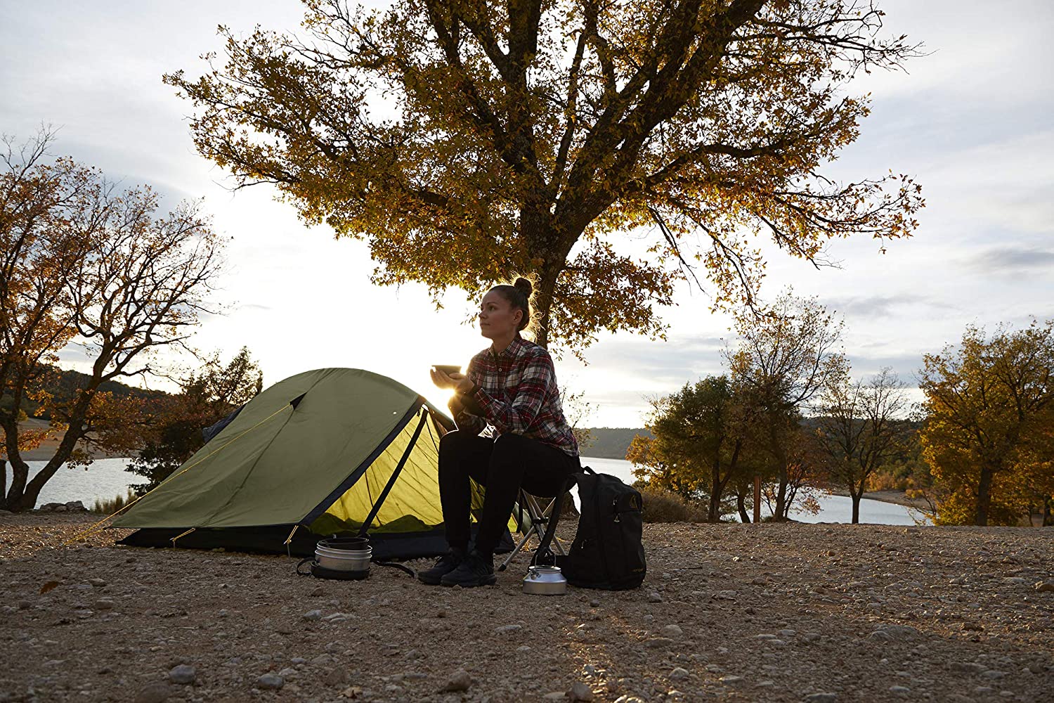 A woman sits by an olive Grand Canyon Cardova 1 tent on a lakeshore, autumn trees behind