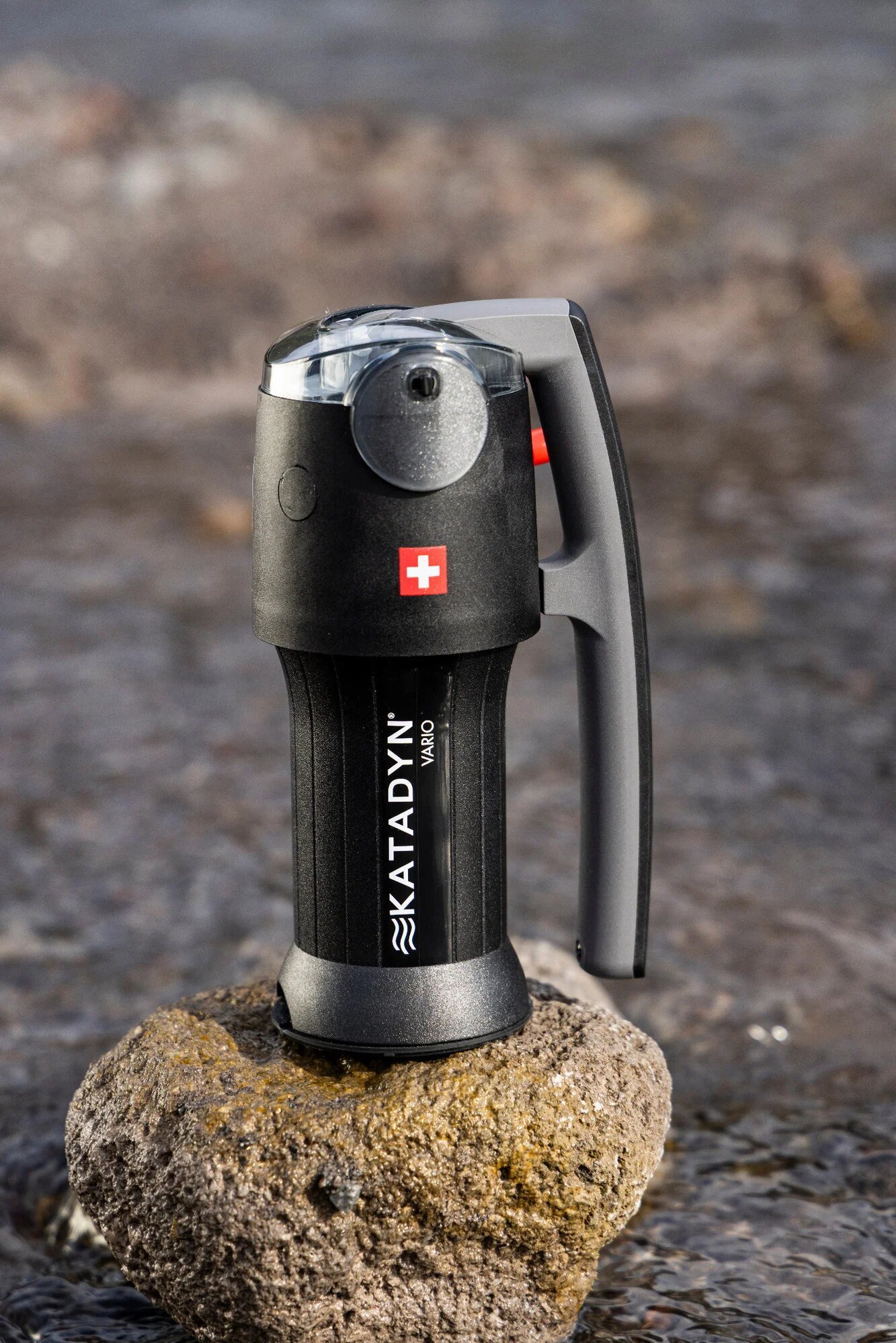 A Katadyn Vario filter standing on a rock by the water, with its black body and a Swiss cross
