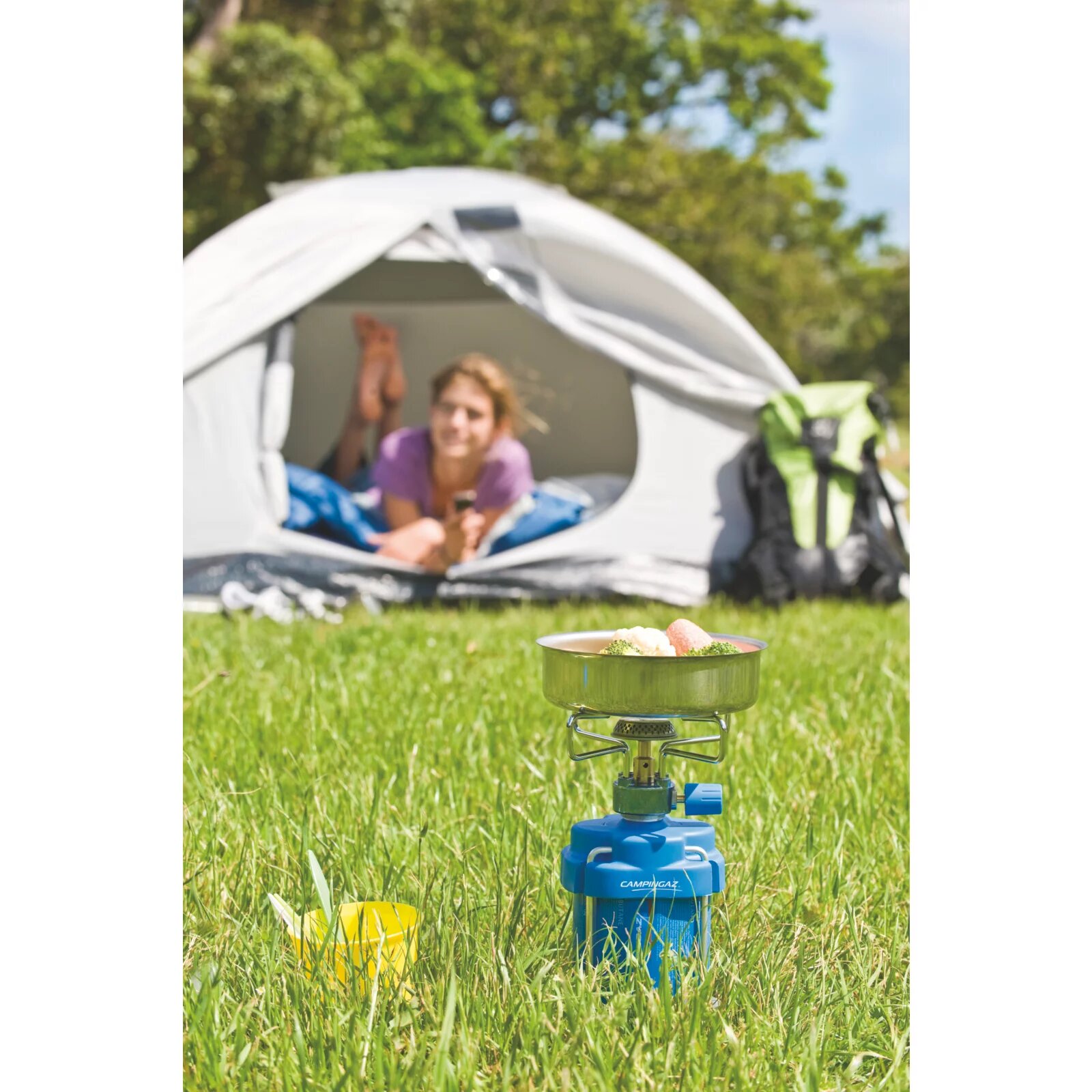 Campingaz Bleuet stove with a pan standing on grass, with a tent and a woman lying in it in the background
