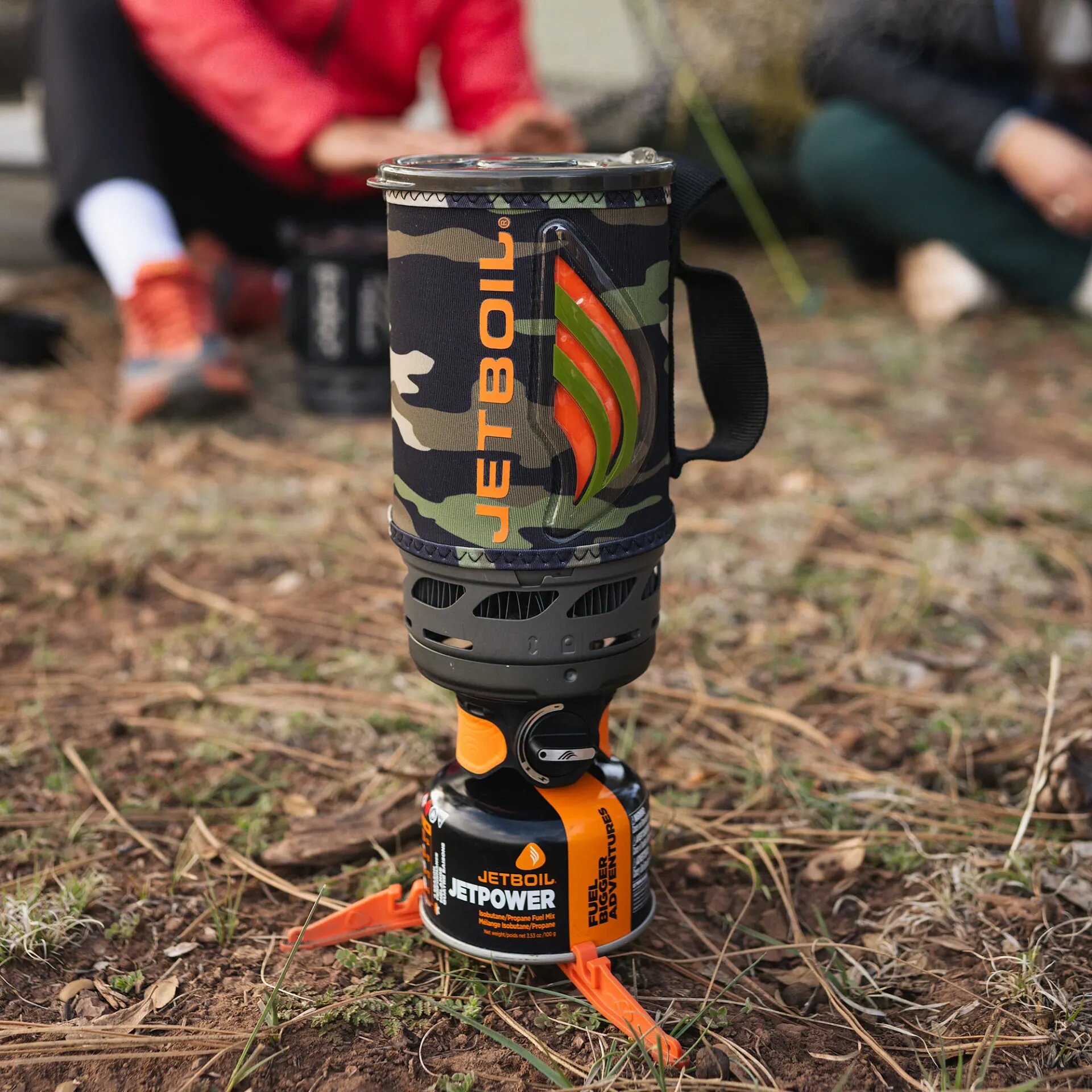 Jetboil Flash 1.0L in camo standing on the ground in a forest on a canister with a tripod base, people sitting in the background