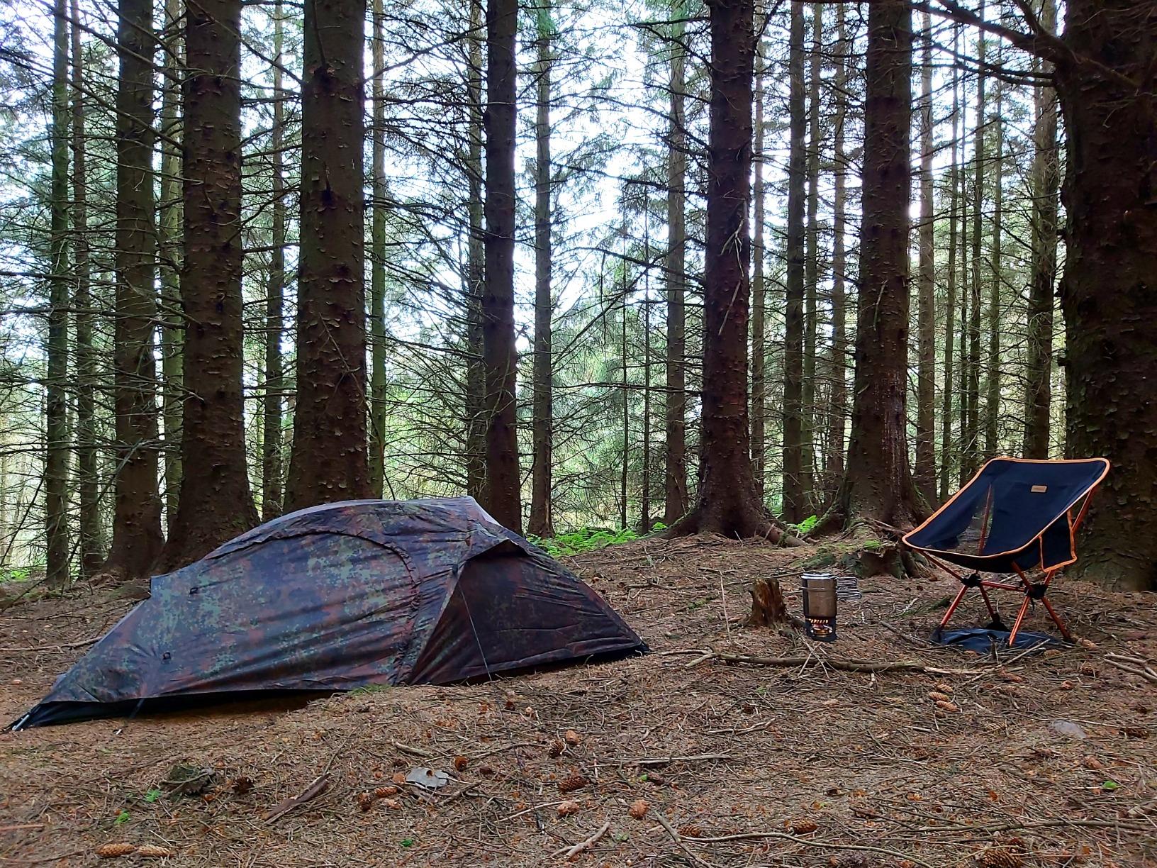 Flecktarn camo tent pitched in a spruce forest, a camping stool and a mug beside it